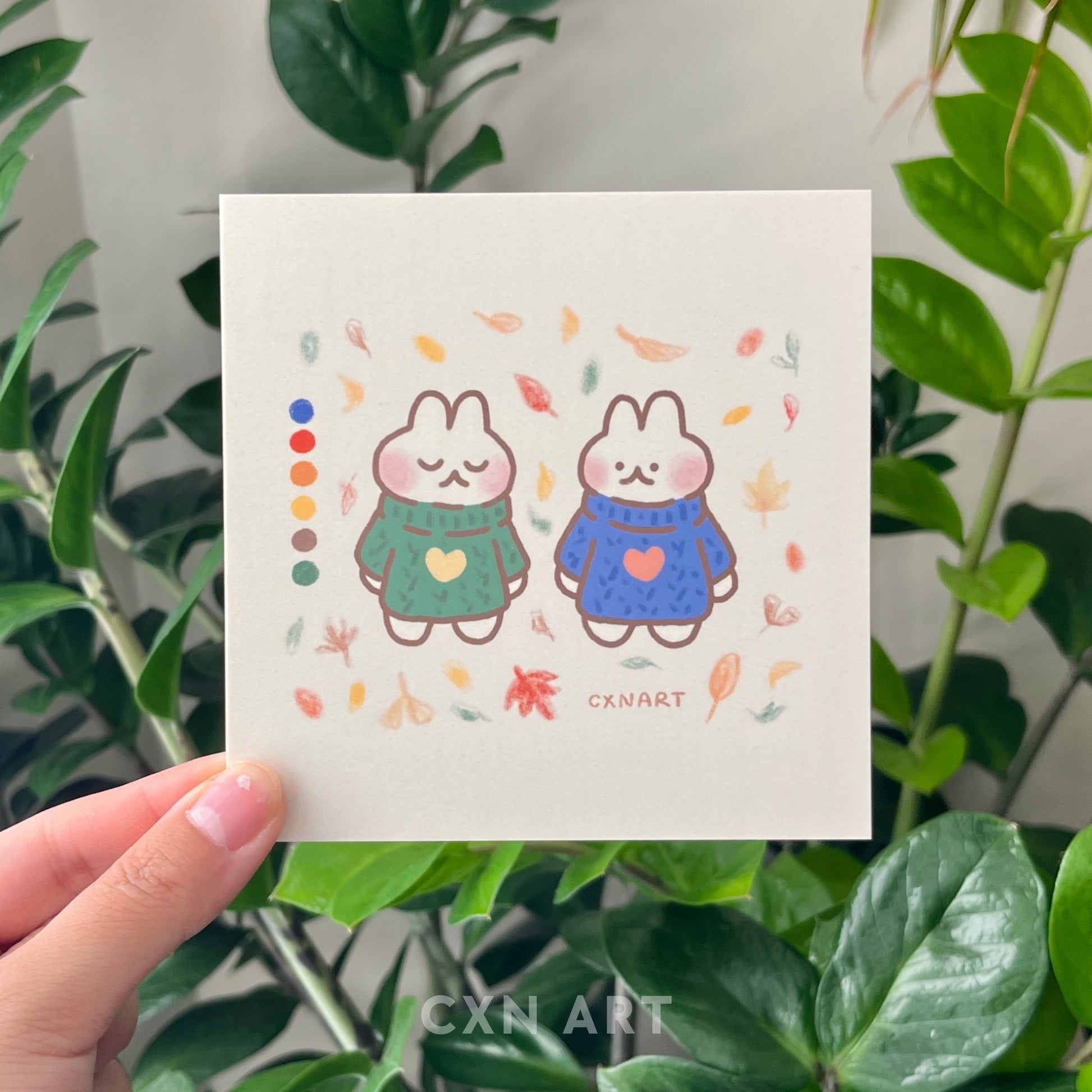 A hand holding a square art print postcard featuring an illustration of two bunnies in autumn colors with leaves around them.