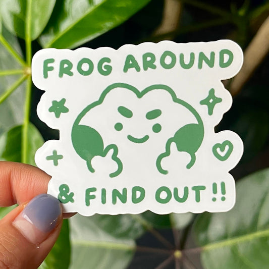 Hand holding a sticker with text "frog around & find out" and a frog holding up two middle fingers with a cheeky smile against a leafy green background