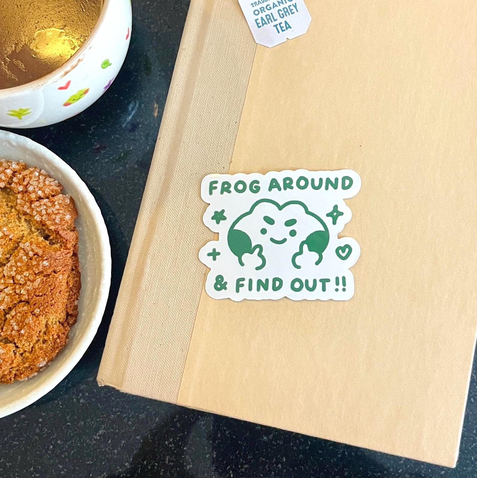 Book with a 'Frog Around & Find Out' sticker on a dark surface with a bowl of food in the background