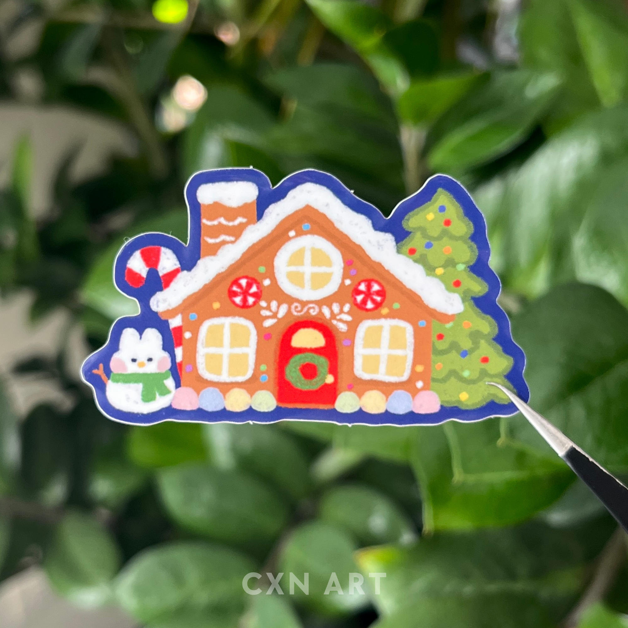 Colorful illustrated gingerbread house sticker with a white bunny character with a green scarf, featuring CXN ART branding. Sticker held against dark, leafy green. 