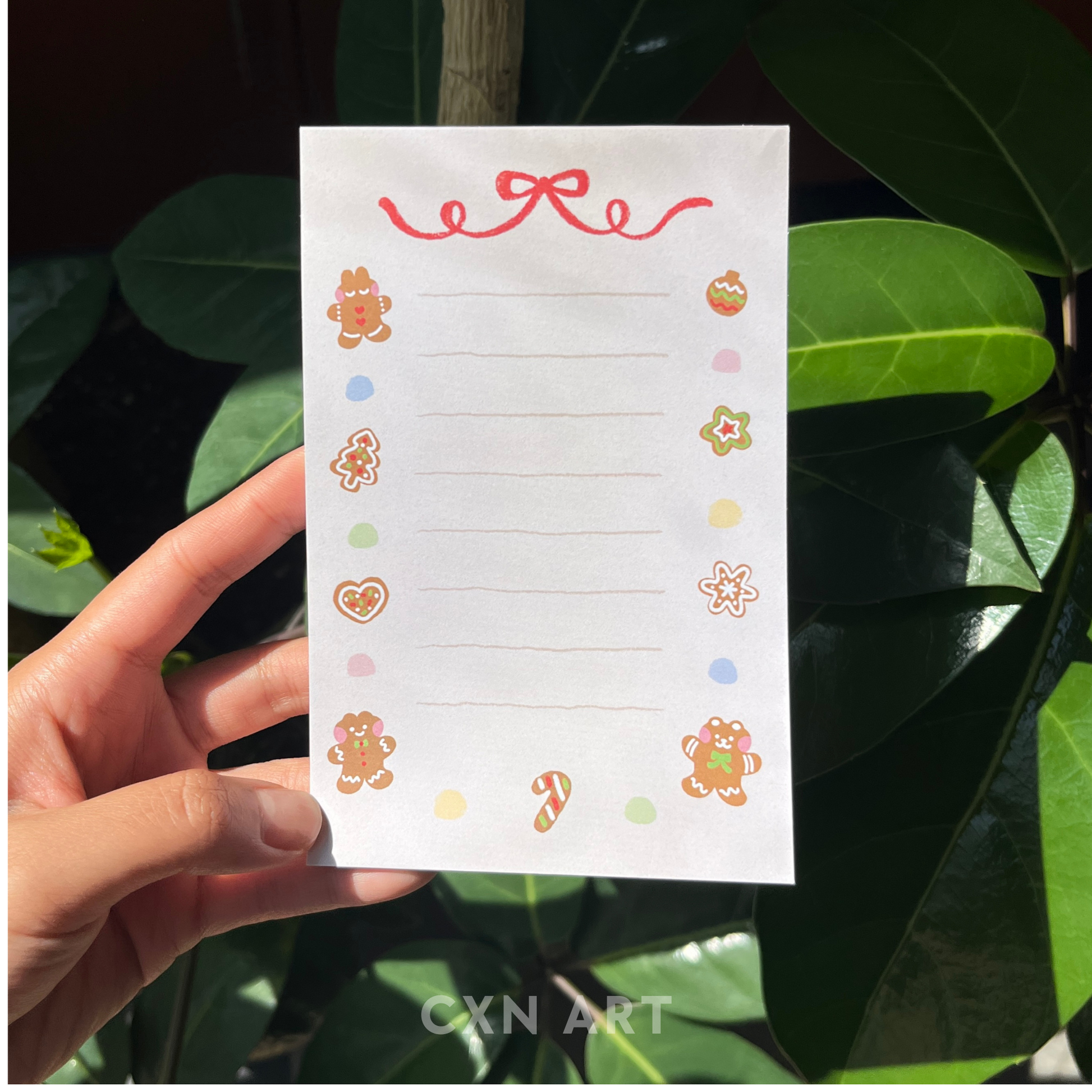 Hand holding a note pad memo pad with decorative gingerbread illustrations against a leafy background