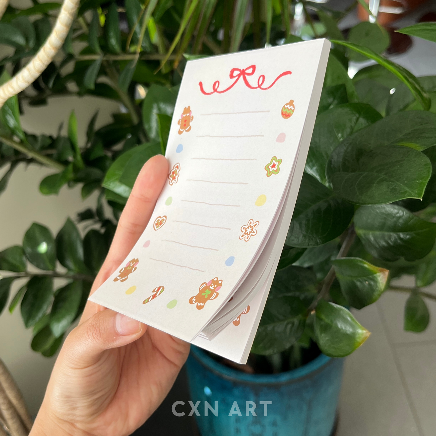 Hand holding a small notepad with gingerbread illustrative designs against a green plant background