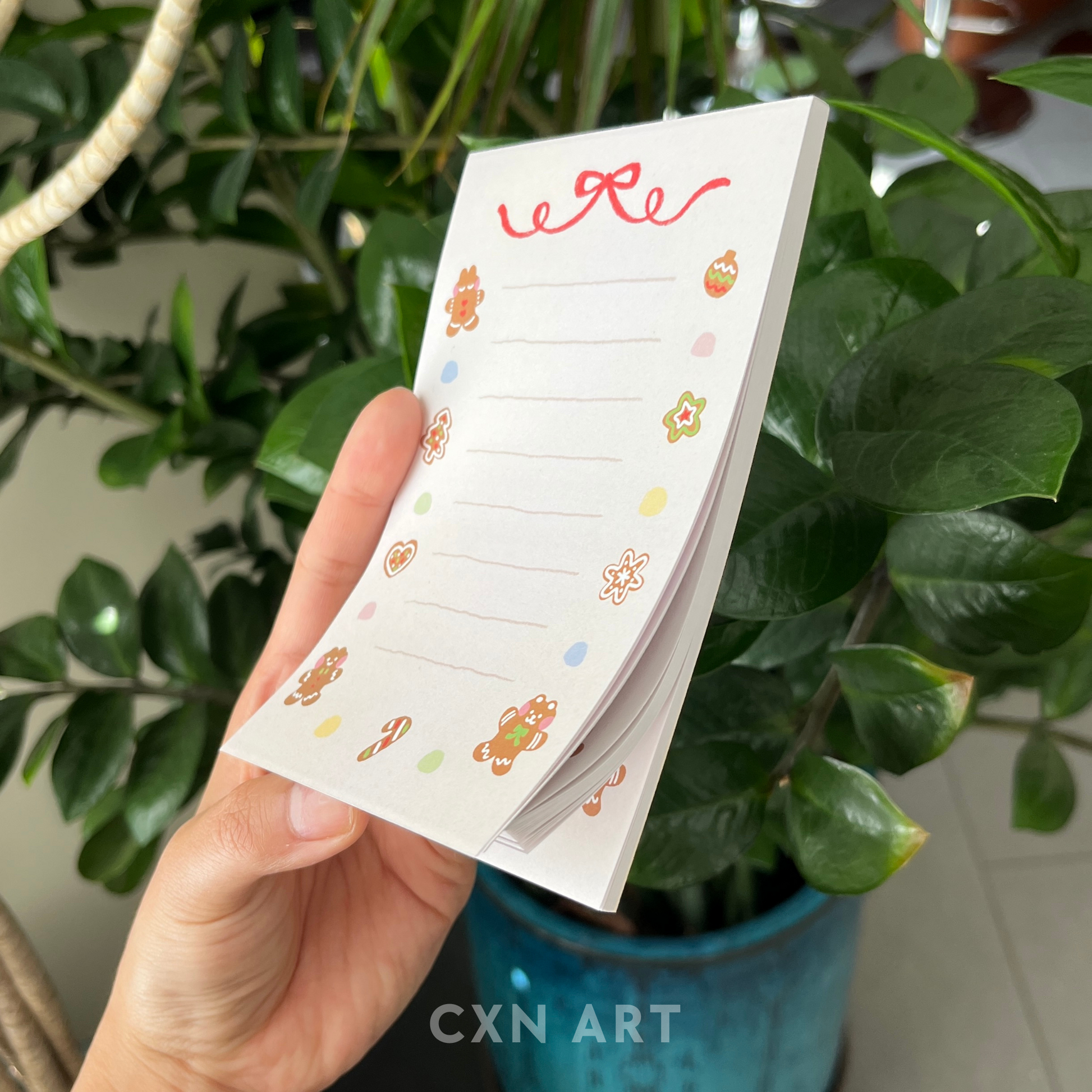 Hand holding a small notepad with gingerbread illustrative designs against a green plant background