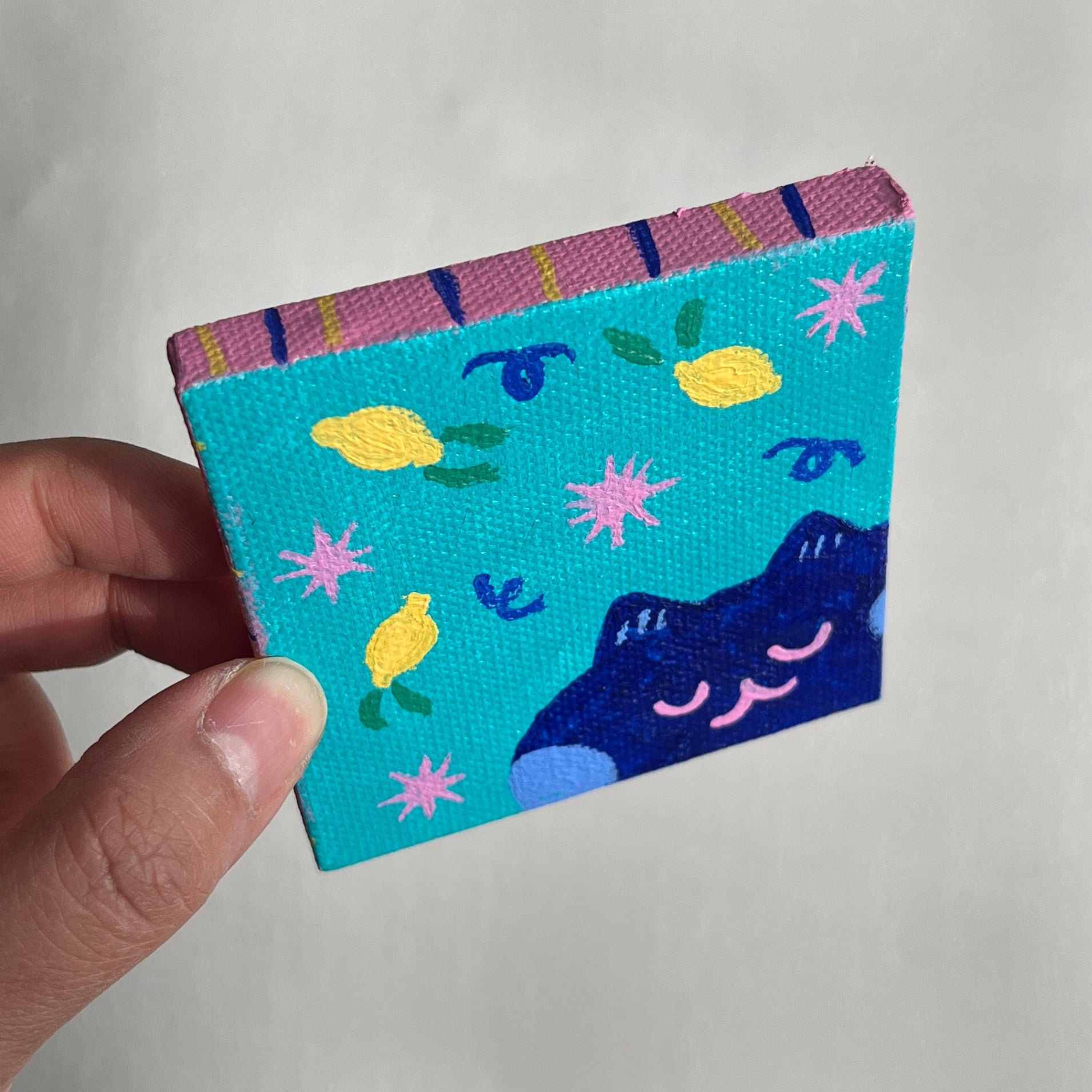 Colorful square painting with whimsy lemon designs, flowers, and cat held by a hand against a light background