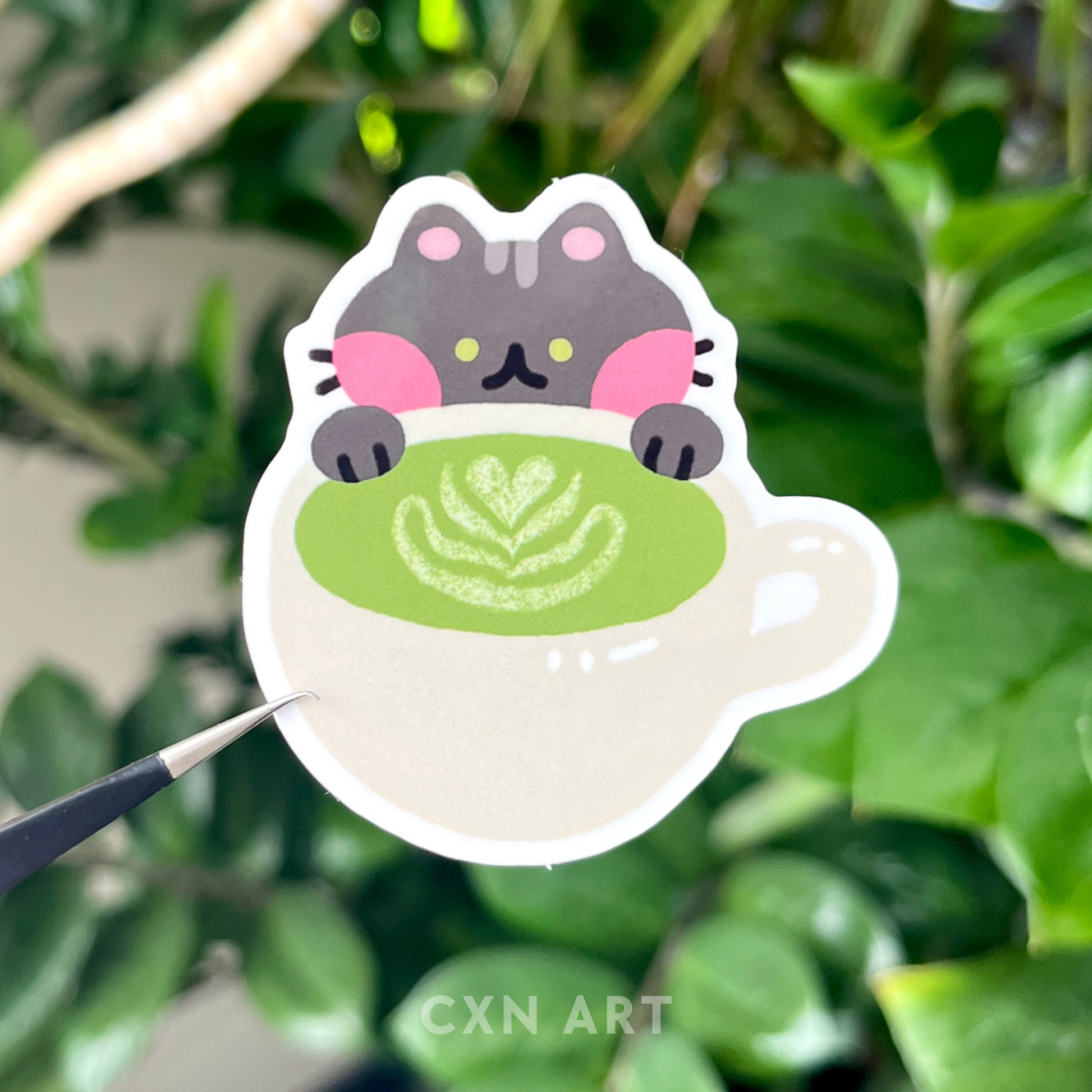 Sticker of a black cat with green eyes leaning against a matcha green tea latte cup, against a green leafy background. Brand 'Cxn Art' visible.
