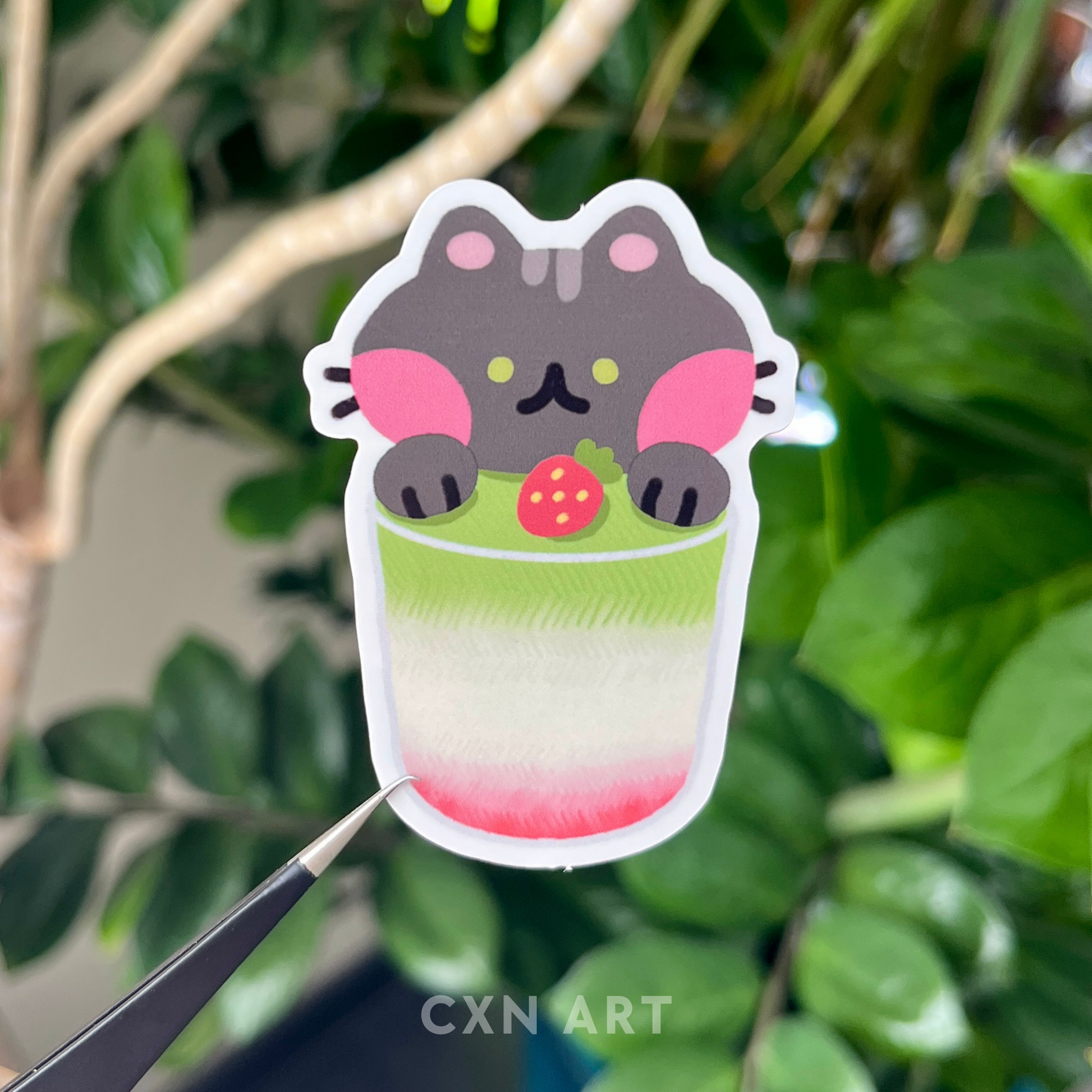 Black cat with green eyes in a cup of strawberry matcha with a strawberry in the drink. Sticker held against a leafy, green background.