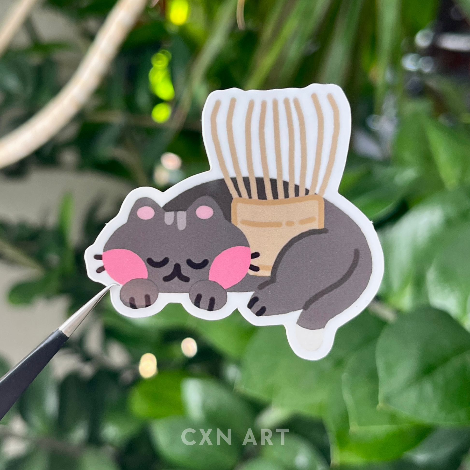 Sticker of a black cat sleeping and holding a bamboo matcha whisk against a green leafy background with 'Cxn Art' branding.