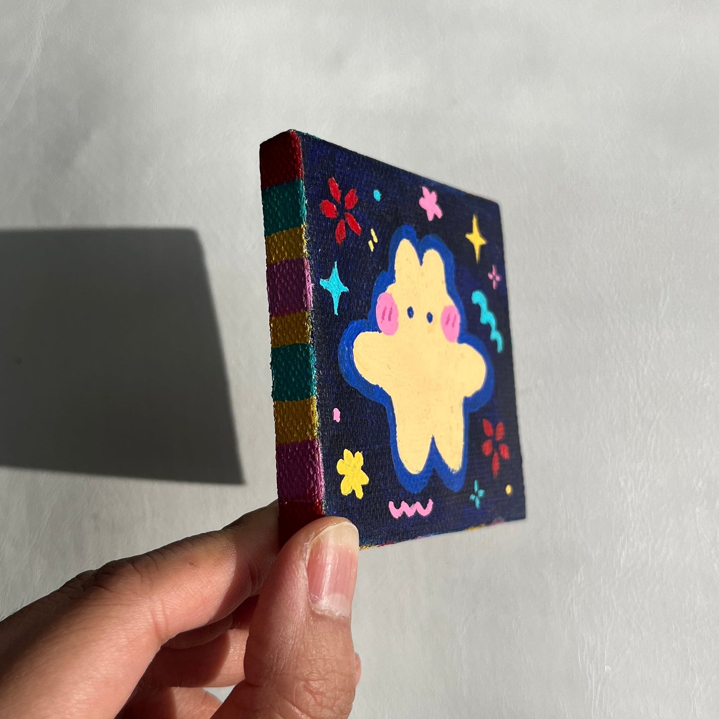 Hand holding a small, colorful paining wih star, charm, flower, and animal design on a plain background