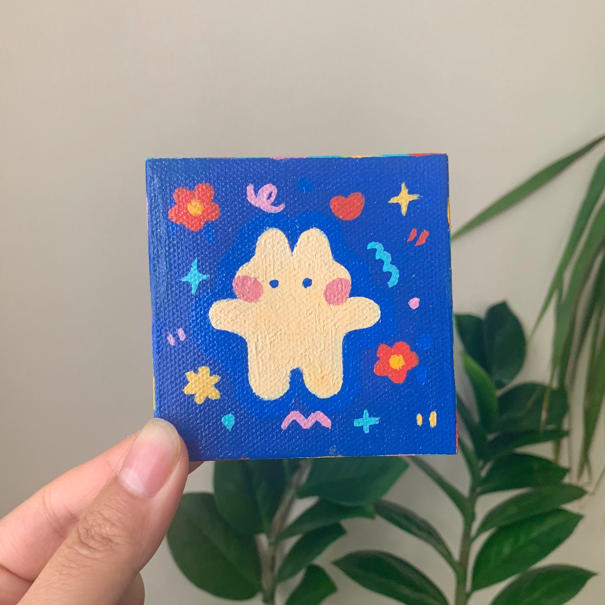 A small acrylic painting of a whimsical bunny design on a dark blue background, held in a person's hand.